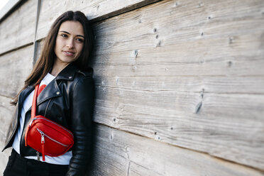 Glückliche junge Frau mit einer roten Hüfttasche, die an einer Holzwand lehnt - JRFF03139