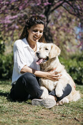 Happy woman sitting on meadow in city park with her Labrador Retriever - JRFF03152