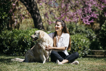 Happy woman sitting on meadow in city park with her Labrador Retriever enjoying sunlight - JRFF03153