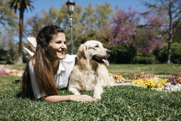 Happy woman lying on meadow in city park with her Labrador Retriever enjoying sunlight - JRFF03154