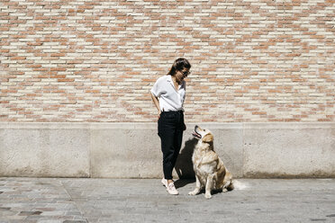 Frau steht vor einer gemusterten Ziegelwand und spricht mit ihrem Labrador Retriever - JRFF03157