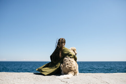 Back view of woman sitting besides her Labrador Retriever at quay looking at horizon - JRFF03162