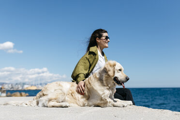 Happy woman sitting besides her Labrador Retriever at quay looking to the sea - JRFF03166