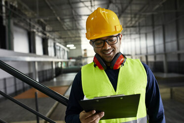 Smiling worker holding clipboard in factory warehouse - ZEDF02254
