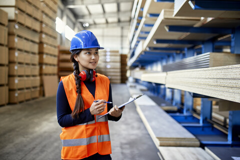 Lächelnde Arbeiterin mit Klemmbrett in einem Fabriklager, lizenzfreies Stockfoto