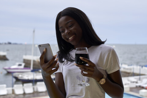 Porträt einer glücklichen jungen Frau mit einem Glas Rotwein, die auf ihr Mobiltelefon schaut, lizenzfreies Stockfoto