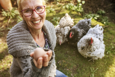Porträt einer glücklichen Frau im Garten mit ihren Orpington-Hennen, die ein Ei zeigen - JATF01138