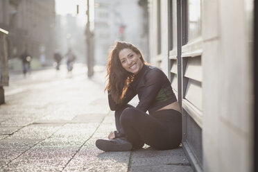 Portrait of happy sportive young woman sitting on the ground in the city - ASCF00991