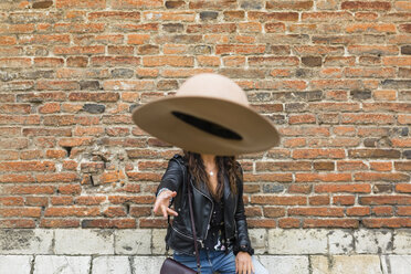 Porträt einer jungen Frau mit schwarzer Lederjacke, Backsteinmauer, werfender Hut - MGIF00390