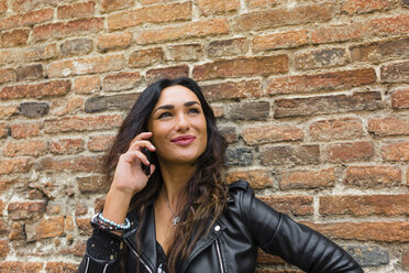 Porträt einer jungen Frau in schwarzer Lederjacke, die ein Smartphone benutzt, mit einer Backsteinmauer im Hintergrund - MGIF00393