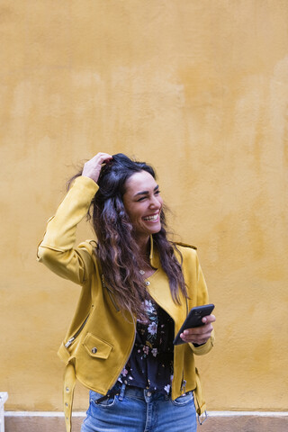 Porträt einer jungen lachenden Frau mit gelber Lederjacke und Smartphone in der Hand, lizenzfreies Stockfoto
