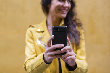 Porträt einer jungen Frau in gelber Lederjacke, die ein Selfie macht, gelbe Wand im Hintergrund - MGIF00405