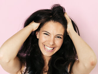 Portrait of young smiling woman with black hair, Hands in hair, in front of pink background - HMEF00363