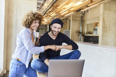 Happy man and woman with laptop fist bumping in modern office - FMKF05634