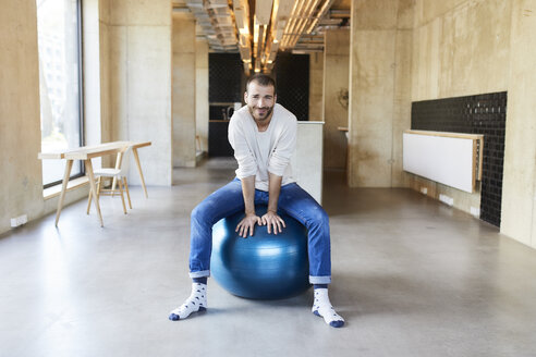 Portrait of young man sitting on fitness ball in modern office - FMKF05646