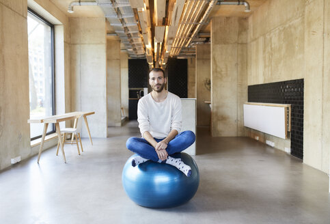 Porträt eines jungen Mannes, der in einem modernen Büro auf einem Fitnessball sitzt - FMKF05647