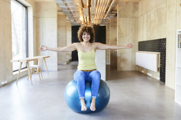 Porträt einer lächelnden Frau, die auf einem Fitnessball in einem modernen Büro sitzt - FMKF05648