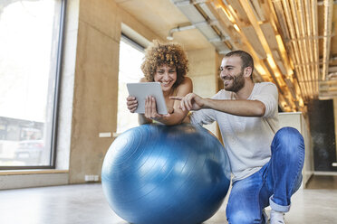 Glücklicher Mann und Frau, die ein Tablet auf einem Fitnessball in einem modernen Büro benutzen - FMKF05650