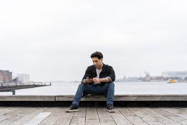 Denmark, Copenhagen, young man sitting at the waterfront using cell phone - AFVF02746