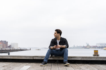 Denmark, Copenhagen, young man sitting at the waterfront with cell phone - AFVF02747