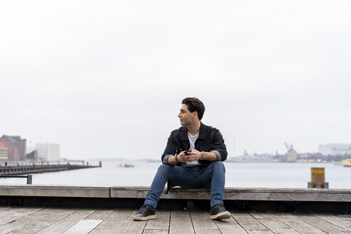 Dänemark, Kopenhagen, junger Mann am Wasser sitzend mit Mobiltelefon - AFVF02747
