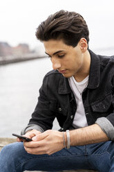 Denmark, Copenhagen, young man sitting at the waterfront using cell phone - AFVF02748