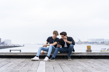 Dänemark, Kopenhagen, zwei junge Männer sitzen am Wasser und telefonieren - AFVF02751
