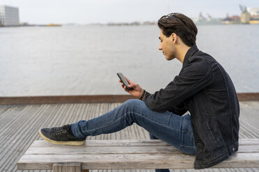 Dänemark, Kopenhagen, junger Mann sitzt auf einer Bank am Wasser und benutzt ein Mobiltelefon - AFVF02777