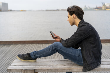 Denmark, Copenhagen, young man sitting on a bench at the waterfront using cell phone - AFVF02777