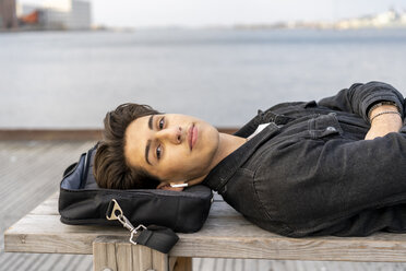 Denmark, Copenhagen, portrait of young man with earbuds lying on a bench at the waterfront - AFVF02789