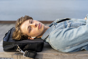 Denmark, Copenhagen, portrait of young man with earbuds lying on a bench at the waterfront - AFVF02793