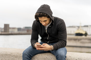 Dänemark, Kopenhagen, junger Mann sitzt auf einer Mauer am Wasser und benutzt ein Mobiltelefon - AFVF02804