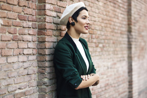 Modische junge Frau mit Baskenmütze und grüner Jacke an einer Backsteinmauer - JSMF01003