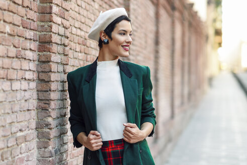 Modische junge Frau mit Baskenmütze und grüner Jacke an einer Backsteinmauer - JSMF01005