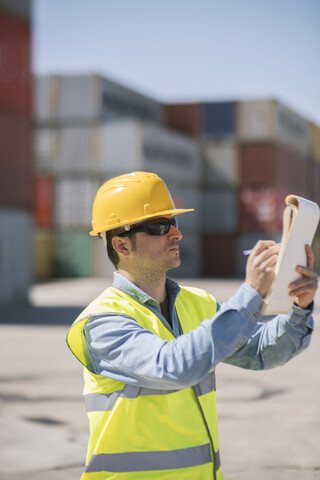 Arbeiter mit einem Notizblock in der Nähe von Frachtcontainern auf einem Industriegelände, lizenzfreies Stockfoto