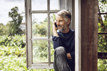 Mann am Handy sitzt am Fenster in tropischer Umgebung - MCF00134