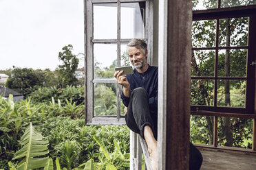 Mann mit Handy sitzt im Fenster in tropischer Umgebung - MCF00153