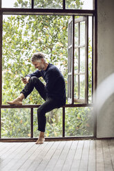 Happy man with cell phone sitting in the window in tropical surrounding - MCF00155