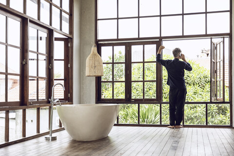 Mann im Badezimmer mit Handy am Fenster in tropischer Umgebung, lizenzfreies Stockfoto