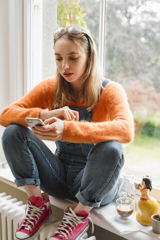 Junge Frau schreibt eine SMS mit einem Smartphone im Fenster, lizenzfreies Stockfoto