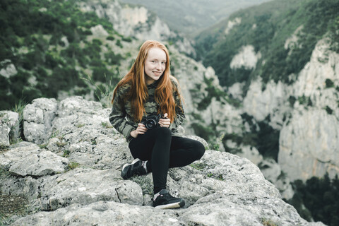 Porträt einer lächelnden kaukasischen Frau, die auf einem Felsen sitzt und eine Kamera hält, lizenzfreies Stockfoto