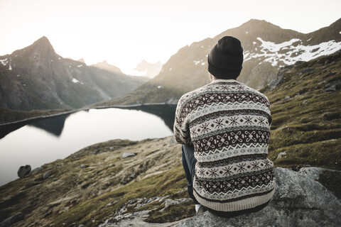 Kaukasischer Mann sitzt auf einem Felsen und bewundert die malerische Aussicht auf einen Bergsee, lizenzfreies Stockfoto