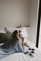 Junge Frau im Bett liegend mit Buch, Sonnenbrille und Polaroidbildern - LHPF00696