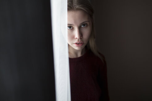Ernste kaukasische Frau steht in der Nähe eines Vorhangs am Fenster - BLEF00265