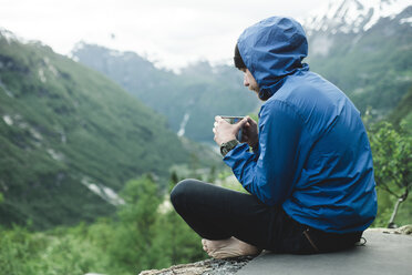 Kaukasischer Mann sitzt in einer Berglandschaft und trinkt Kaffee - BLEF00553