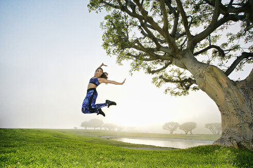 Gemischtrassige Frauen springen auf einem Feld in der Nähe eines Baumes - BLEF00662