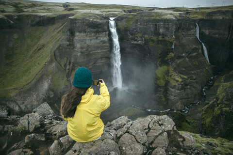 Kaukasische Frau sitzt auf einer Klippe und fotografiert einen Wasserfall, lizenzfreies Stockfoto