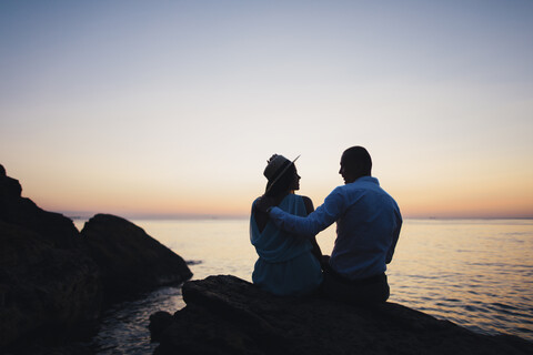 Silhouette von kaukasischen Paar sitzt auf einem Felsen in der Nähe von Meer bei Sonnenuntergang, lizenzfreies Stockfoto