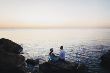 Kaukasisches Paar sitzt bei Sonnenuntergang auf einem Felsen am Meer - BLEF00933