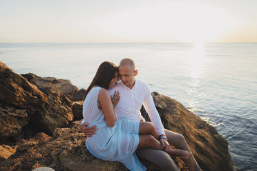 Caucasian couple hugging on rocks on beach at sunset - BLEF01040